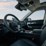 Jaecoo 7 SUV interieur vanaf bestuurderszijde met zwarte leren stoelen, digitaal dashboard display, stuurwiel met Jaecoo-branding, centraal touchscreen en panoramadak met bewolkte lucht zichtbaar