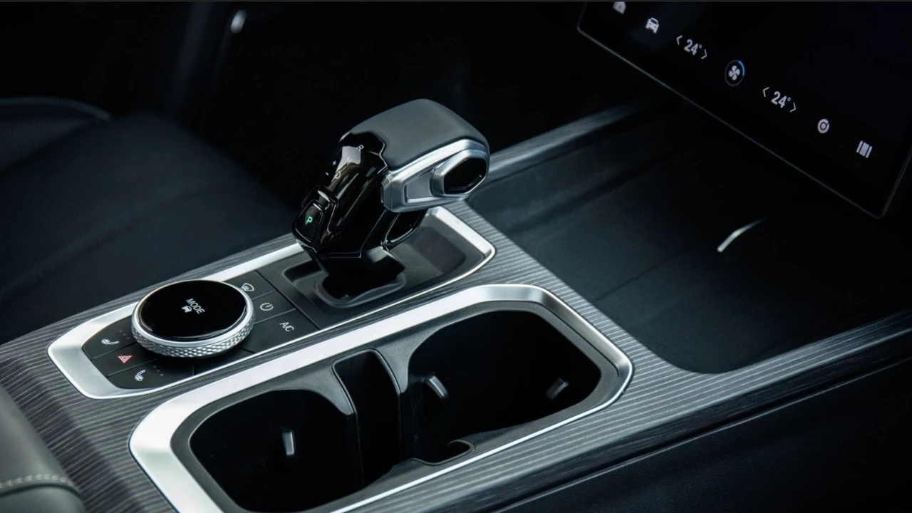 Jaecoo 7 SUV middenconsole met moderne versnellingspook, roterende bedieningsknop, dubbele bekerhouders, koolstofvezel-stijl trim, leren stoelen en dashboard touchscreen zichtbaar op achtergrond