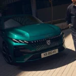 Close-up vooraanzicht van de 2025 Peugeot 308 facelift in teal groen met drieklauw LED signatuurverlichting en verlicht embleem met persoon die in de buurt loopt voor schaalverhouding