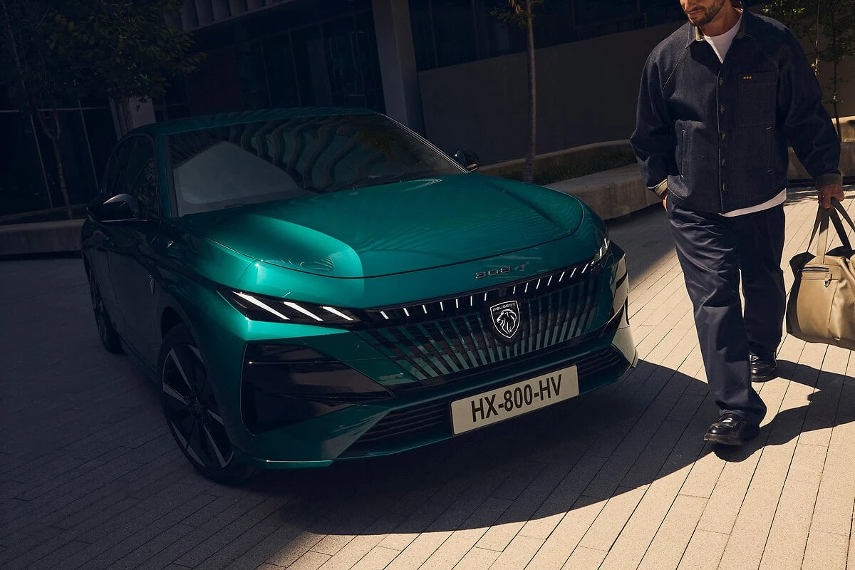 Close-up vooraanzicht van de 2025 Peugeot 308 facelift in teal groen met drieklauw LED signatuurverlichting en verlicht embleem met persoon die in de buurt loopt voor schaalverhouding