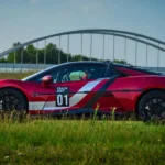 Rode Yangwang U9 Track Edition elektrische supercar met Track Test 01 livery gepositioneerd op gras met moderne brugarchitectuur op de achtergrond bij Duitse testfaciliteit