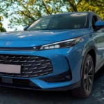 2025 MG HS Hybrid+ SUV in Arctic Blue kleur geparkeerd op bomenlaan, vooraanzicht drie-kwart hoek met LED-koplampen, zwarte honingraat grille en 19-inch lichtmetalen velgen