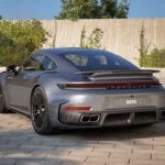 Zilveren 2025 Porsche 911 Turbo S vanuit achterste driekwart hoek in buitenomgeving met betonnen muur achtergrond, achterbumper en brede carrosserie ontwerp
