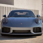 Frontale vooraanzicht van zilveren 2025 Porsche 911 Turbo S in buitenomgeving met brede stance, grote voor luchtinlaten, LED koplampen en moderne architecturale achtergrond