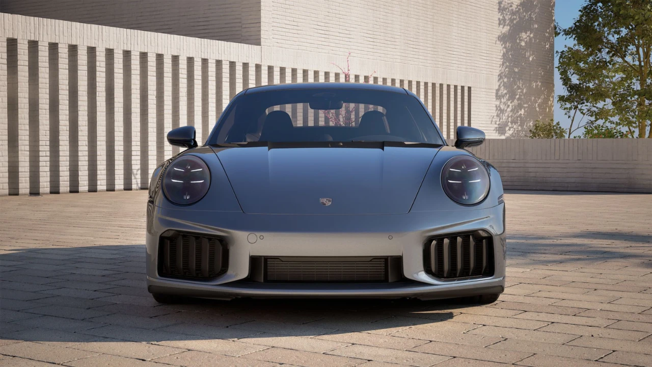 Frontale vooraanzicht van zilveren 2025 Porsche 911 Turbo S in buitenomgeving met brede stance, grote voor luchtinlaten, LED koplampen en moderne architecturale achtergrond