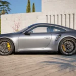 Volledig zijprofiel van zilveren 2025 Porsche 911 Turbo S in buitenomgeving met klassieke 911 proporties, gele remklauwen, zij luchtinlaten en meerspaaks lichtmetalen velgen