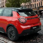 Rode 2026 Nissan Micra elektrische auto vanuit achter driekwart aanzicht op kasseienstraat met ronde achterlichten, Nissan-branding en sportieve achterspoiler