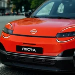 Lage hoek aanzicht van rode 2026 Nissan Micra op stadsstraat met nadruk op frontontwerp met ronde LED-koplampen en moderne bumperstyling