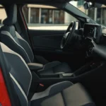 Zijaanzicht van 2026 Nissan Micra voorstoelen met sportieve zijsteunen, grijs en wit contrastmateriaal, en rood deurpaneel zichtbaar