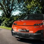 De 2026 Nissan Micra elektrische supermini in levendige oranje kleur, met zijn compacte afmetingen van 3,97 meter lengte, moderne LED-verlichting en 18-inch wielen op een schilderachtige landweg.