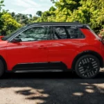 Zijprofiel aanzicht van rode 2026 Nissan Micra met zwart dak met volledig carrosserieontwerp, kenmerkende zijsculptuur en 18-inch wielen in parkdecor