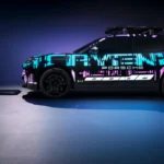 Zijaanzicht van 2026 Porsche Cayenne Electric prototype met cyaan en paarse digitale camouflage livery gepositioneerd boven draadloze oplaadpad, met PORSCHE branding en dakaccessoires