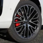 Close-up van 2026 Audi Q3 20-inch zwart lichtmetalen wiel met machinaal gepolijste spaak gezichten met ingewikkeld veelspaaks Y-ontwerp patroon, rode remklauw met Audi branding zichtbaar door spaken, lageprofiels band en wit carrosseriepaneel erboven