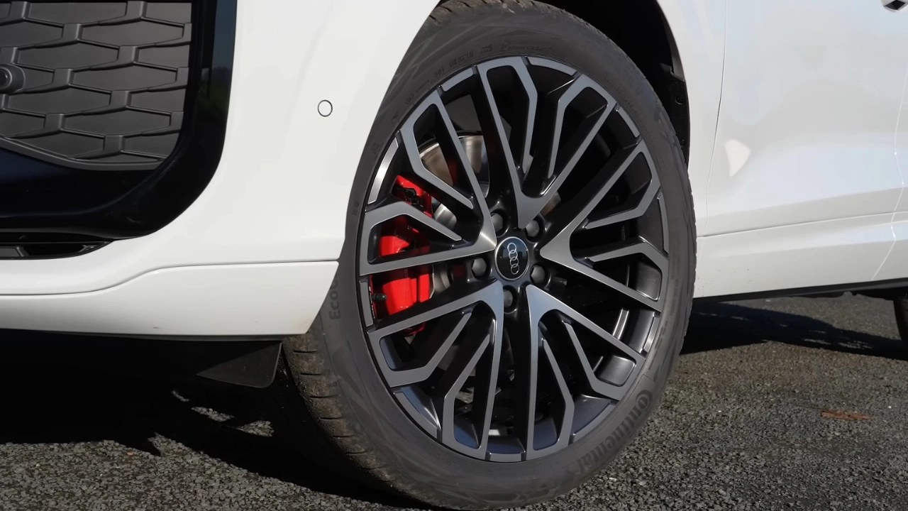 Close-up van 2026 Audi Q3 20-inch zwart lichtmetalen wiel met machinaal gepolijste spaak gezichten met ingewikkeld veelspaaks Y-ontwerp patroon, rode remklauw met Audi branding zichtbaar door spaken, lageprofiels band en wit carrosseriepaneel erboven