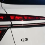 Verlichte 2026 Audi Q3 achterlicht met rode LED-segmenten in bovenste laag die pijlachtig patroon creëren, dunne horizontale rode lichtstrook verbindend naar midden, onderste LED-sectie met verticale geribbelde details en Q3-badge op wit zijpaneel