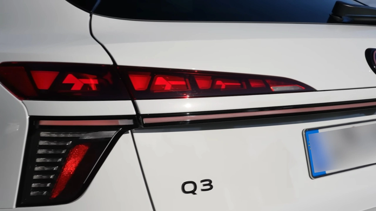 Verlichte 2026 Audi Q3 achterlicht met rode LED-segmenten in bovenste laag die pijlachtig patroon creëren, dunne horizontale rode lichtstrook verbindend naar midden, onderste LED-sectie met verticale geribbelde details en Q3-badge op wit zijpaneel