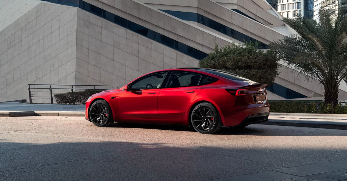 Red Tesla electric sedan in side profile view parked on pavement next to modern gray building with palm trees, featuring black wheels and distinctive fastback roofline