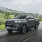 2026 Toyota Hilux BEV in Ash Grey rijdend op een natte weg, vooraanzicht driekwart met brandende LED-koplampen.