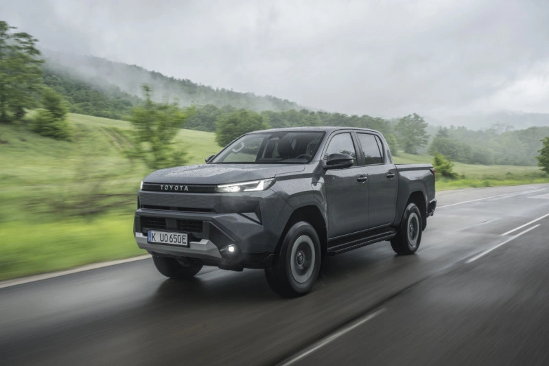 2026 Toyota Hilux BEV in Ash Grey rijdend op een natte weg, vooraanzicht driekwart met brandende LED-koplampen.