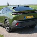 Schuin achteraanzicht van de groene 2026 Peugeot 408 Plug-in Hybrid bij een weiland.