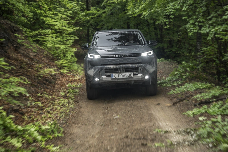 2026 Toyota Hilux BEV op een modderig bospad, vooraanzicht met brandende koplampen, die de off-road kwaliteiten toont.