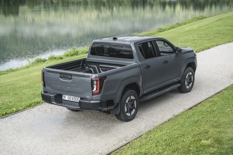 Achteraanzicht driekwart van de 2026 Toyota Hilux BEV in Ash Grey aan een meer, met zicht op de open laadbak.