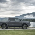Zijaanzicht van de 2026 Toyota Hilux BEV in Ash Grey, geparkeerd aan een meer met bergen op de achtergrond.