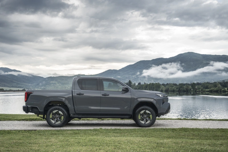 Zijaanzicht van de 2026 Toyota Hilux BEV in Ash Grey, geparkeerd aan een meer met bergen op de achtergrond.